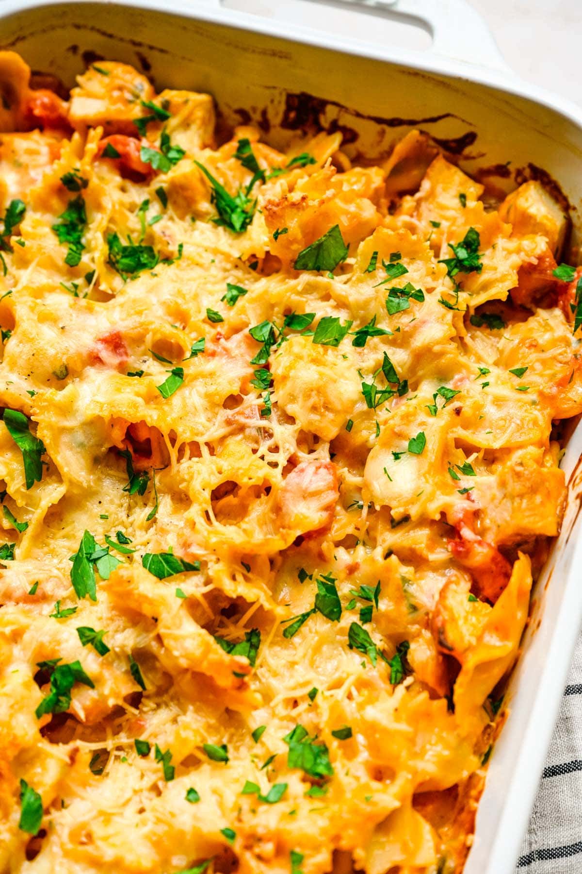A close up photo of a chicken pasta bake in a white rectangular casserole dish, highlighting the melted cheese topping and garnished with parsley.