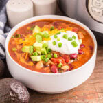 Instant Pot Turkey Chili in a white bowl, garnished with sour cream, shredded cheese, diced avocado and sliced green onion.