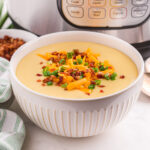 Instant Pot Potato Soup in a decorative white bowl garnished with shredded cheese, bacon and green onion, sitting in front of an Instant Pot.