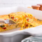 A slice of Cottage Cheese Egg Bake being lifted out of a white casserole dish by a silver serving spatula with a wooden handle, with melted cheese pulling from the rest of the casserole.