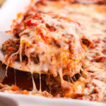 A serving of Ravioli Lasagna being lifted from a white casserole dish, highlighting the melted mozzarella layers.