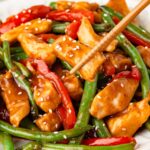 A piece of sauce-covered chicken being picked up by brown chopsticks from a plateful of Chicken and Green Bean Stir Fry.