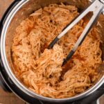 Shredded Buffalo Chicken being taken from an Instant Pot with a pair of black and silver kitchen tongs, viewed from a high angle.