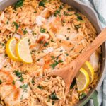 Golden seared chicken breast nestled into creamy lemon orzo garnished with lemon slices and fresh parsley, in a green and grey skillet, being scooped by a wooden spoon.