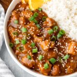 A closeup of Instant Pot Orange Chicken served on a rimmed white plate with a side of white rice, topped with sesame seeds, sliced green onion and orange slices.