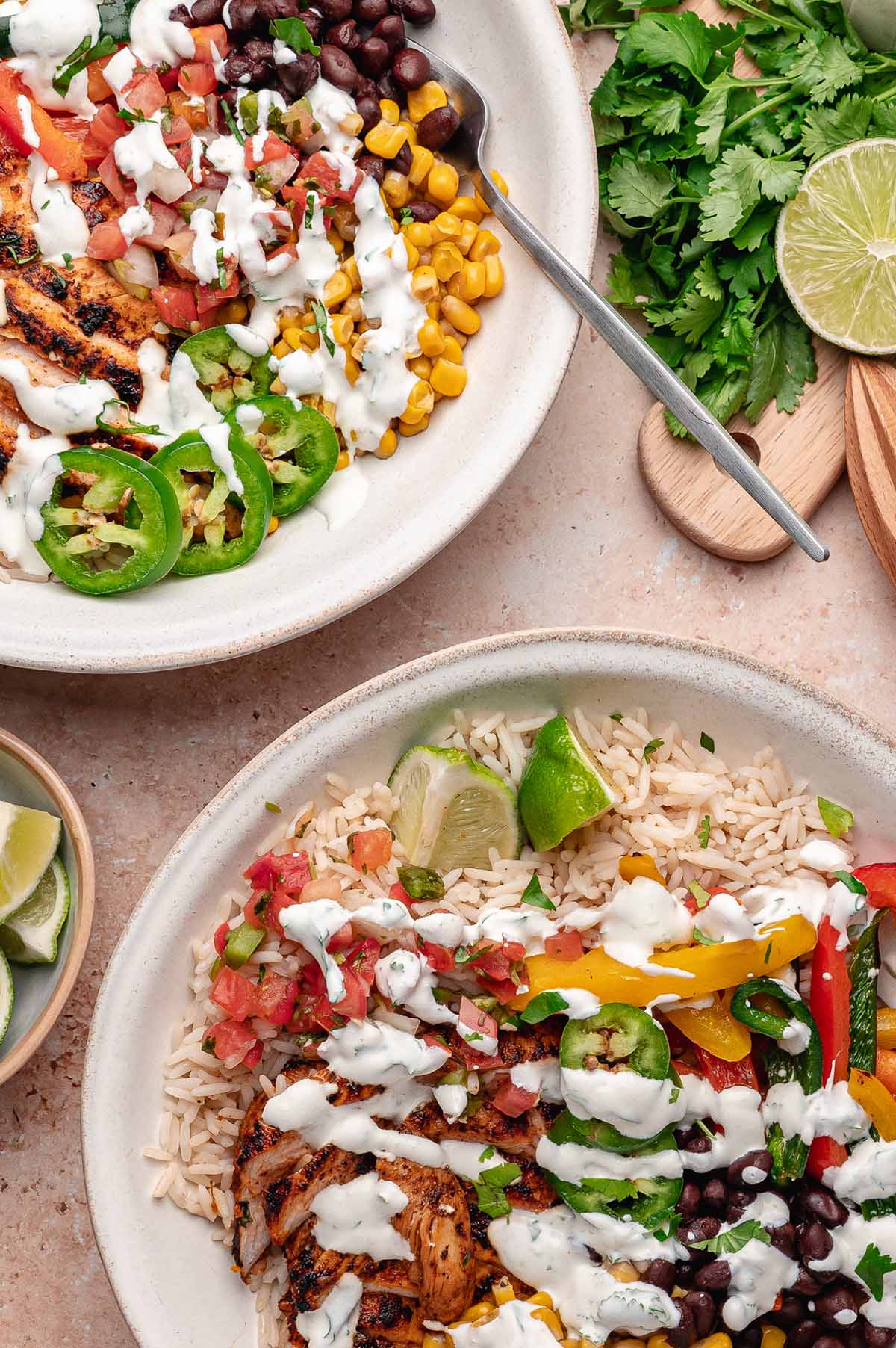 Two chicken fajita bowls with crema, jalapeños, corn, black beans, and lime wedges on a tan-coloured surface.
