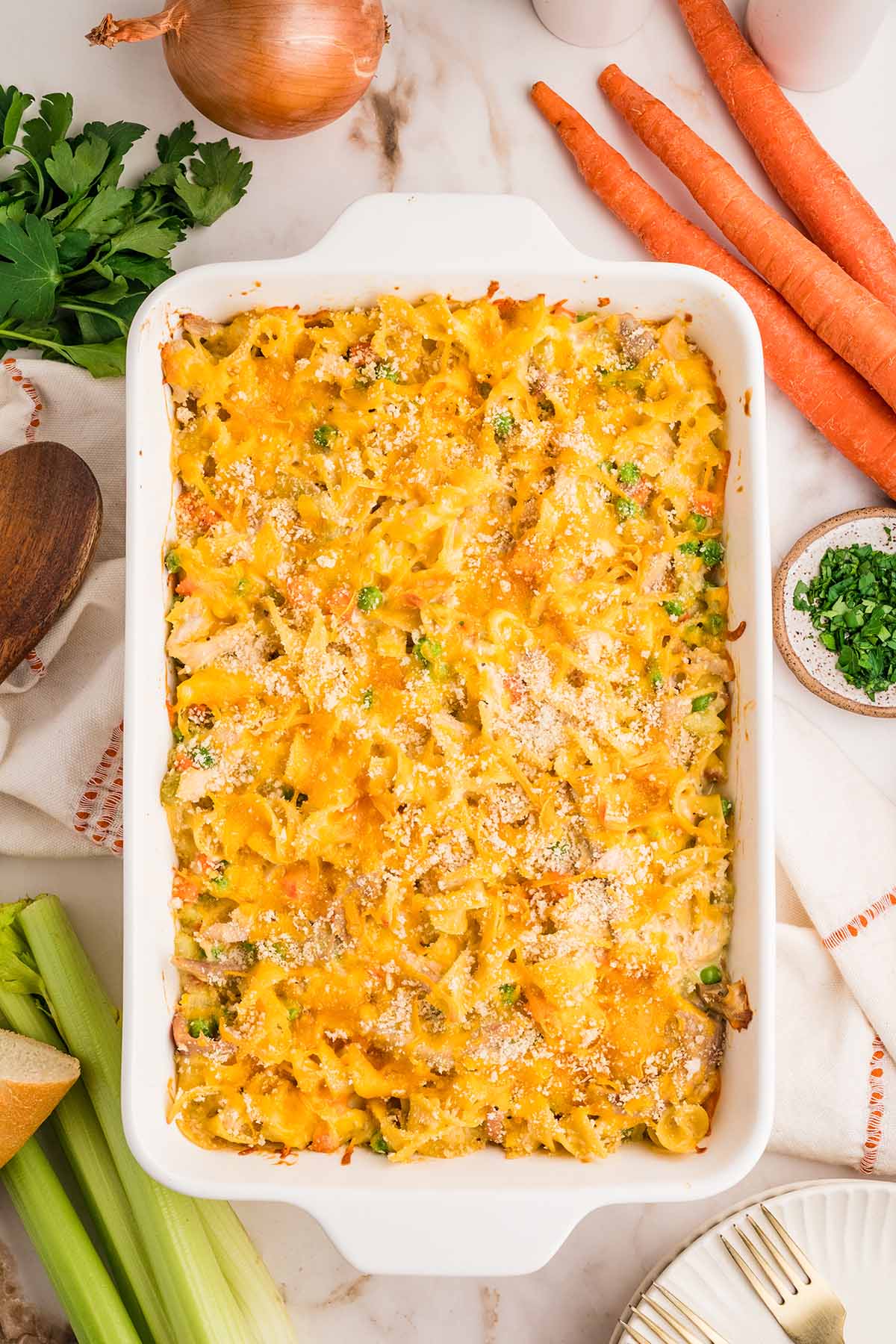 Baked turkey noodle casserole in a white dish surrounded by carrots, celery, onion, and fresh parsley.